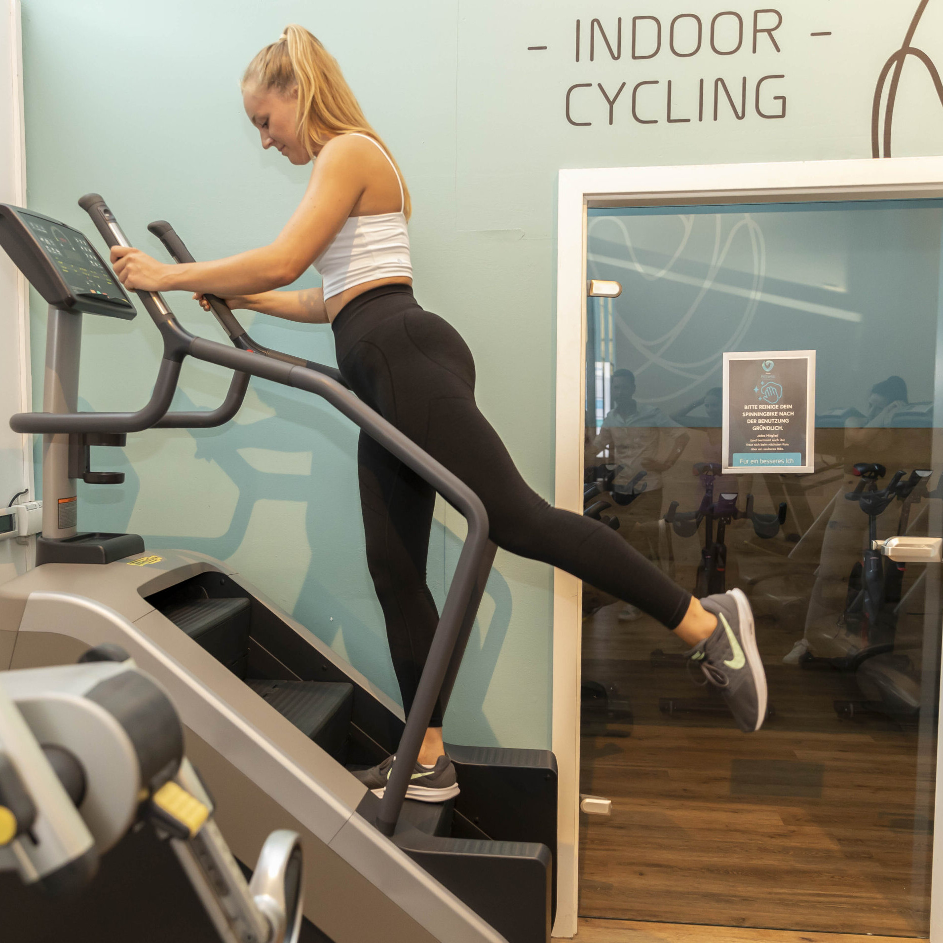 Eine sportliche Frau läuft auf einem Stepper (oder auch Klapptritt genannt) und hält sich aktiv auf der abwärts bewegenden Treppe. Sie schaut auf das Display des Fitnessgeräts. Hintergrund ist eine Glastür, deren Spiegelung weitere Personen zeigt, die auf Standfahrrädern (auch bekannt als Spin Bikes) ihre Kondition trainieren.