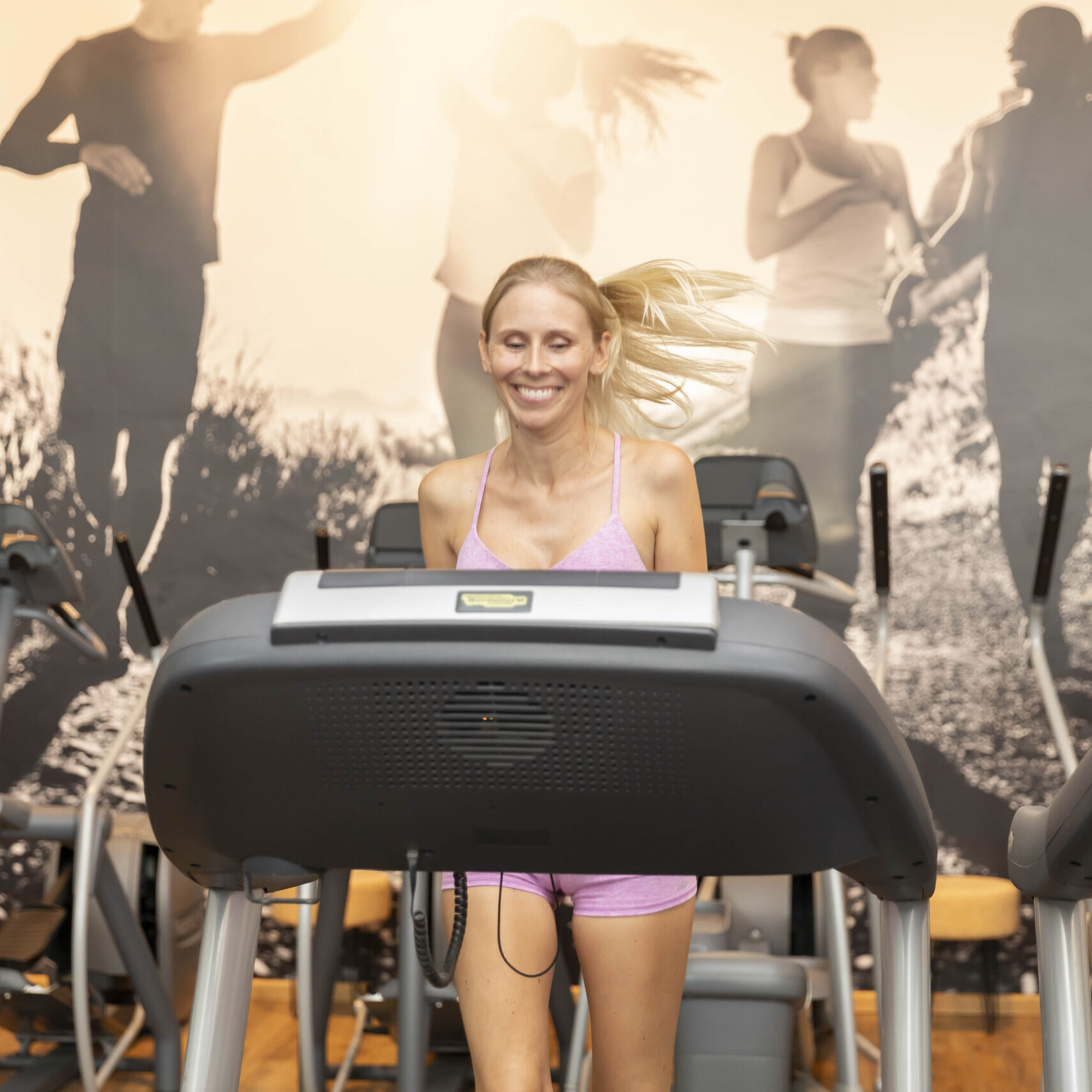 Eine blonde Frau in pinker Trainingskleidung joggt mit einem strahlenden Lächeln auf einem Laufband. Sie befindet sich in einem Fitnessstudio, und an der Wand im Hintergrund hängt ein großes Bild von Sportlern, die im Sonnenschein ihrer sportlichen Aktivität nachgehen.