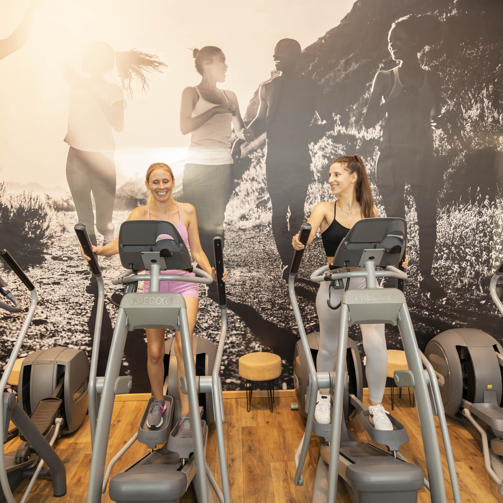 Zwei Frauen trainieren auf Crosstrainern in einem Fitnessstudio. Beide lächeln und unterhalten sich, während sie nebeneinander auf PRECOR-Crosstrainern trainieren. Im Hintergrund ist eine große Wand mit einem Schwarz-Weiß-Foto von Menschen beim Joggen in der Natur zu sehen.