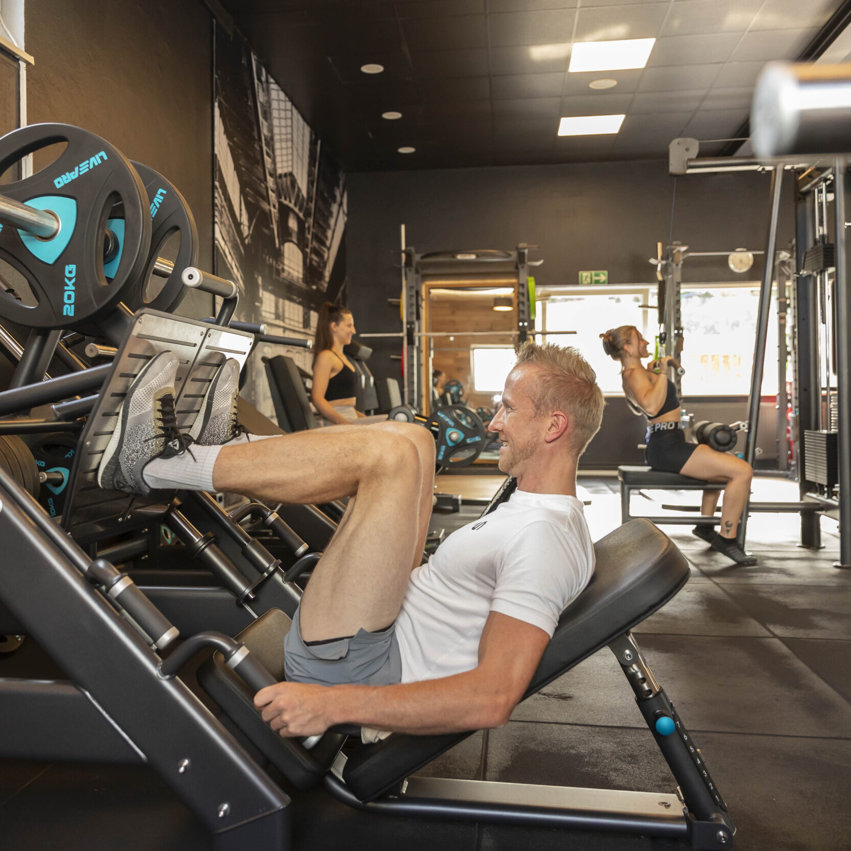 Ein muskulöser Mann liegt motiviert und glücklich auf einer Beinpresse und trainiert intensiv. Im Hintergrund sind zwei Frauen, die in einem gut ausgestatteten Fitnessstudio ihrem Training nachgehen.